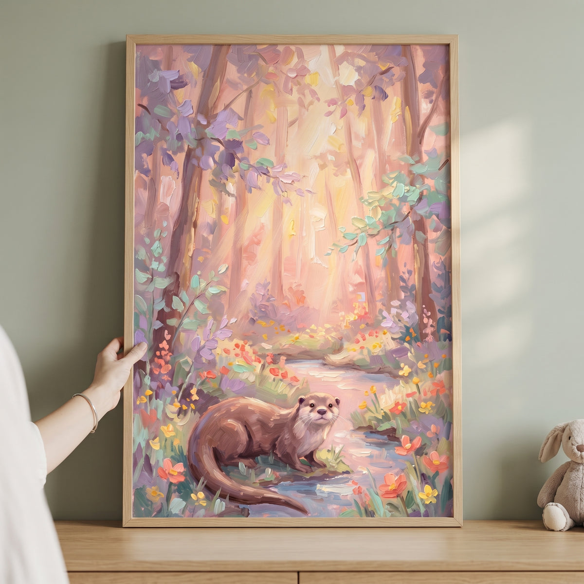 Hand holds framed pastel stream otter woodland wall art of an otter in a forest by a stream, next to a plush bunny.
