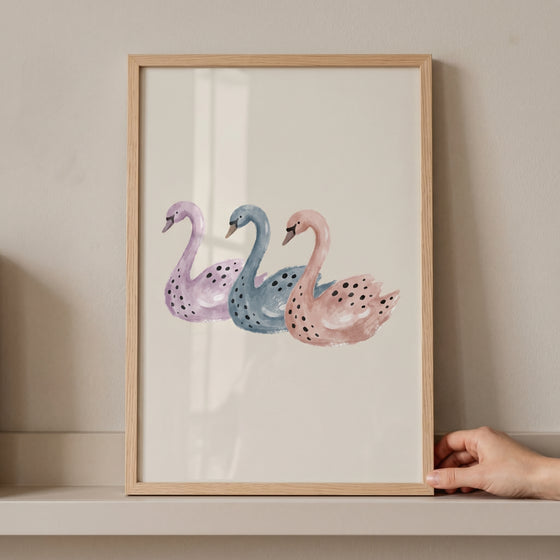 Three purple, blue, and pink swans in a wooden frame. This pastel watercolor spotted swan wall art sits on a shelf.
