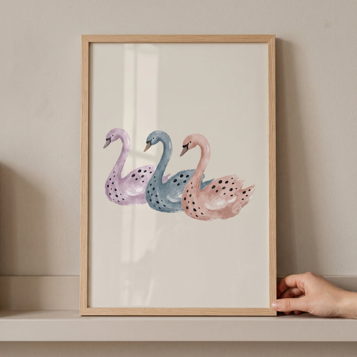 Three purple, blue, and pink swans in a wooden frame. This pastel watercolor spotted swan wall art sits on a shelf.