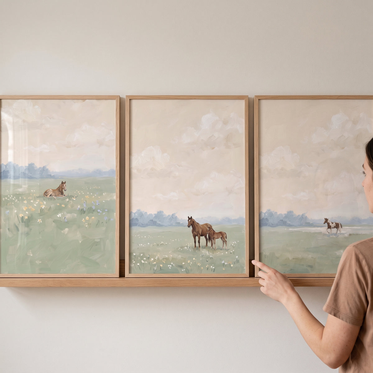 A pastoral sage meadow horses wall art set of 3 in light wood frames rests on a shelf next to a woman's hand and shoulder.