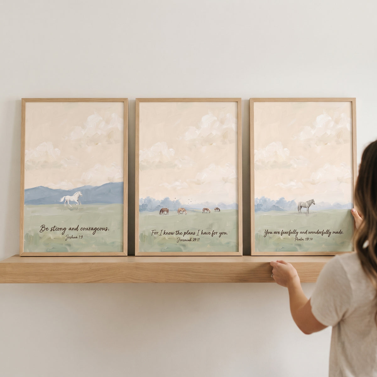 Woman's hands adjust a pastoral scripture meadow horses wall art set of 3 featuring Bible verses on a wooden shelf.