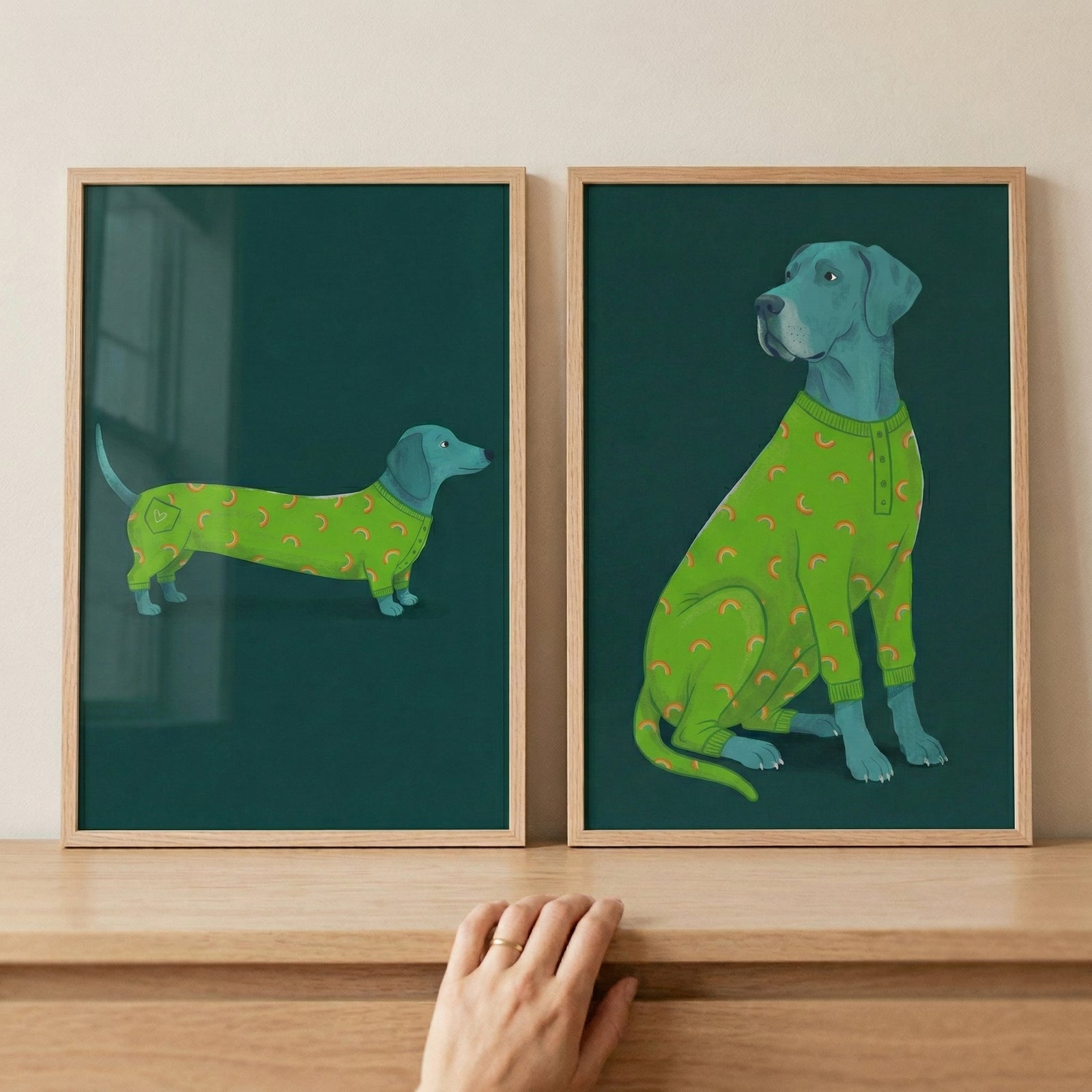 A pair of framed digital illustrations displayed side-by-side on a light-toned wooden surface. Both pieces feature a dark forest green background and a blue-grey dog wearing a vibrant lime green one-piece garment adorned with small orange patterns. 
