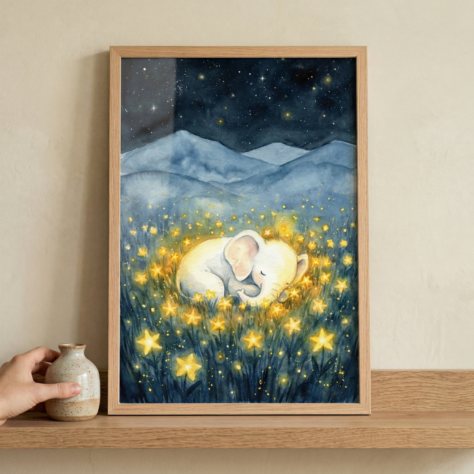A whimsical watercolor-style illustration of a small, white elephant sleeping peacefully in a field of glowing, yellow stars. In the background, soft blue mountain ranges rest under a dark night sky filled with shimmering stars. The scene has a magical, serene atmosphere. The artwork is presented in a light wooden frame, displayed on a wooden shelf alongside a small, speckled ceramic jar. The soft brushstrokes and celestial theme create a dreamlike quality.