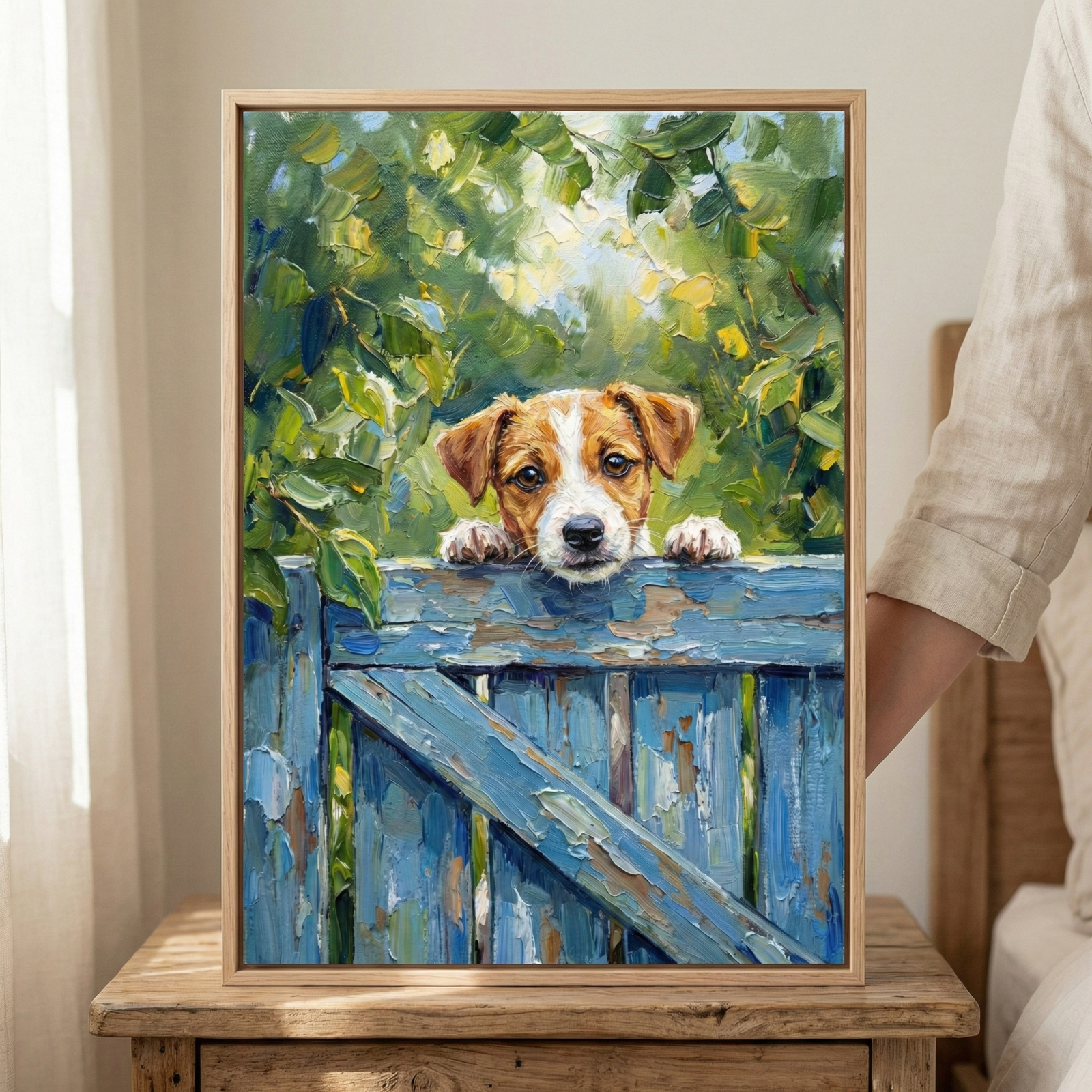 An impressionistic artwork featuring a small brown and white puppy peeking over the top of a weathered blue wooden gate. Its large brown eyes and front paws rest on the textured rail. The background is filled with lush green foliage and dappled sunlight, rendered with thick, visible brushstrokes. The piece is held in a light wood frame, resting on a rustic wooden side table in a brightly lit room next to a white curtain.