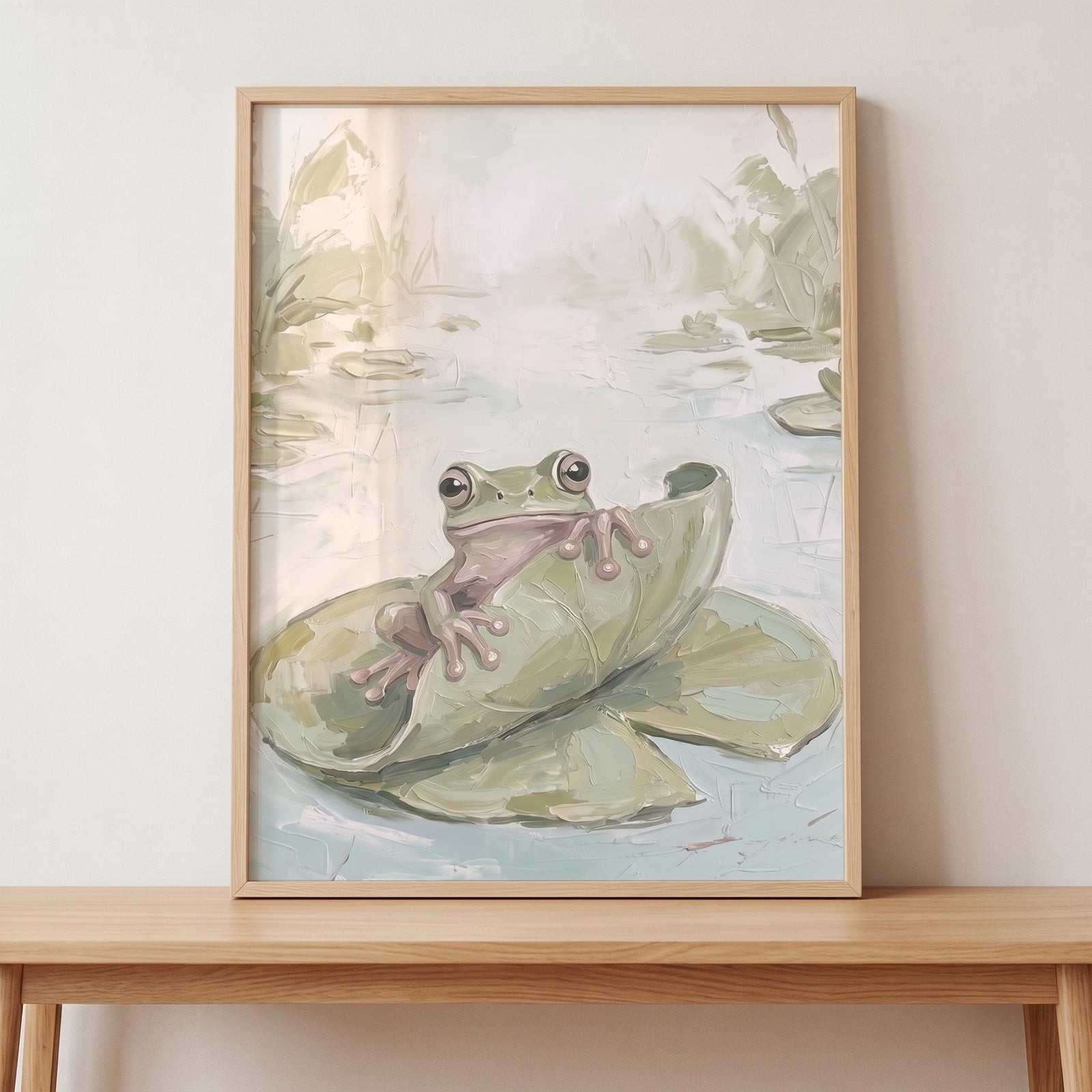 Peeking leaf green frog wall art shows a textured frog peering over a lily pad, framed on a wooden table.