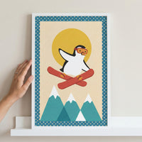 A hand touches a white-framed illustration of a cute cartoon penguin wearing orange goggles and skiing through the air. The artwork features a large yellow sun and three stylized teal mountains with snowy peaks on a light background. The print has a blue border with white polka dots and is displayed on a white shelf.