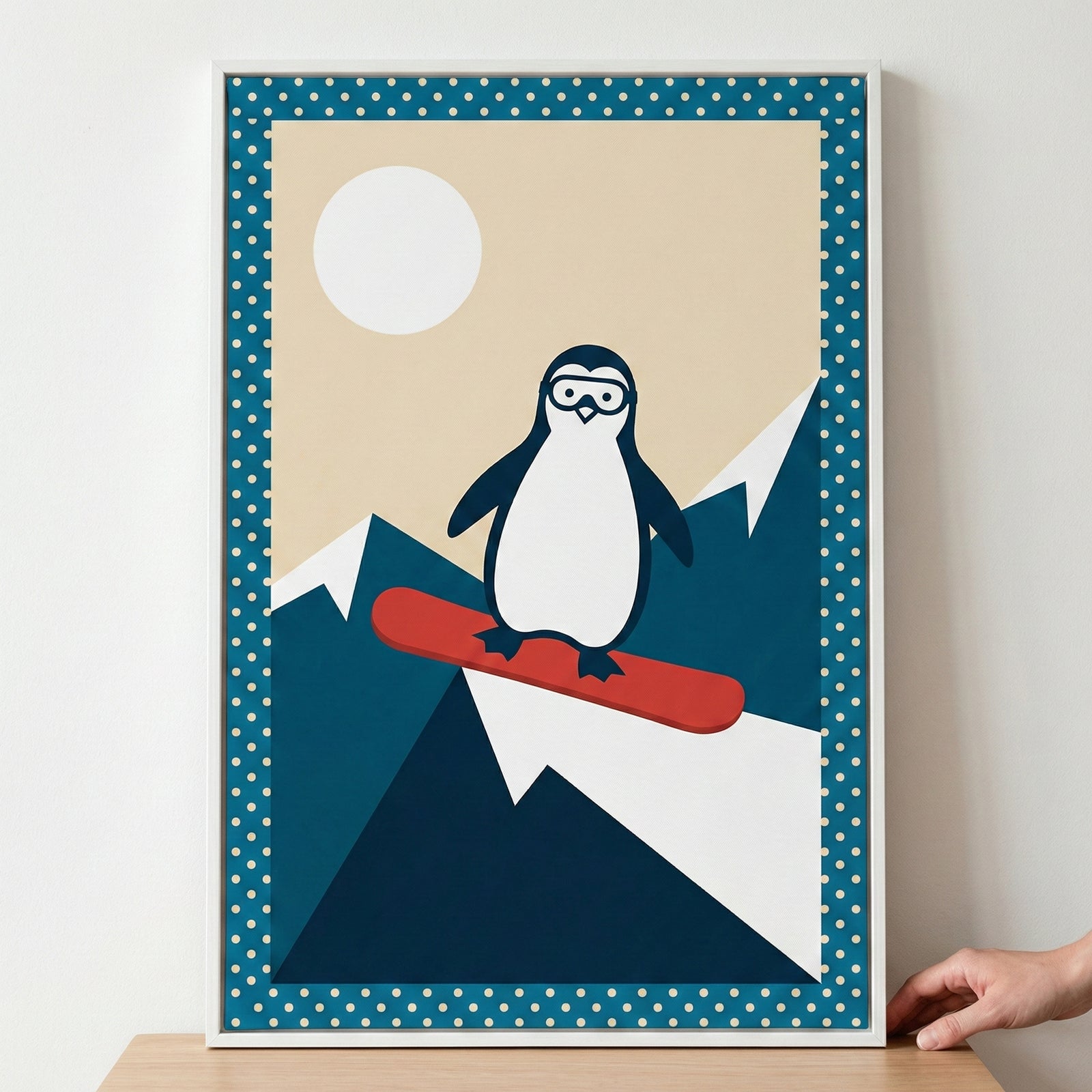 A minimalist graphic illustration of a cute cartoon penguin wearing goggles and riding a red snowboard. The penguin is positioned in the center, set against a background of stylized dark blue and white mountain peaks. Above the mountains is a pale beige sky featuring a large white circular sun or moon. The entire illustration is enclosed within a decorative dark blue border featuring small white polka dots and is presented in a simple white frame resting on a wooden surface.