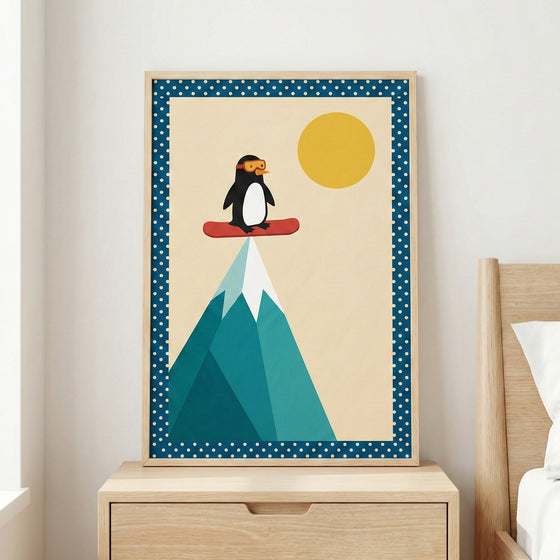A framed minimalist illustration sits on a light wooden nightstand next to a bed. The artwork features a cute cartoon penguin wearing orange goggles, balancing on a red snowboard at the very tip of a stylized, geometric mountain. The mountain is composed of several shades of teal and blue triangles with a white snowy peak. A large, solid yellow sun is positioned in the cream-colored sky. The image has a blue border with white polka dots and is housed in a simple light wood frame.