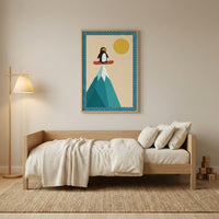 A cozy, minimalist room features a light wood daybed dressed in ivory bedding and soft pillows. To the left stands a wooden floor lamp with a cream-colored shade. Centered on the wall above the bed is a framed illustration of a cartoon penguin standing on a red board at the summit of a geometric teal mountain peak. The artwork includes a large yellow sun against a cream background, all framed by a blue polka-dotted border. The room is completed by a textured tan rug on the floor and a small stack of wooden 