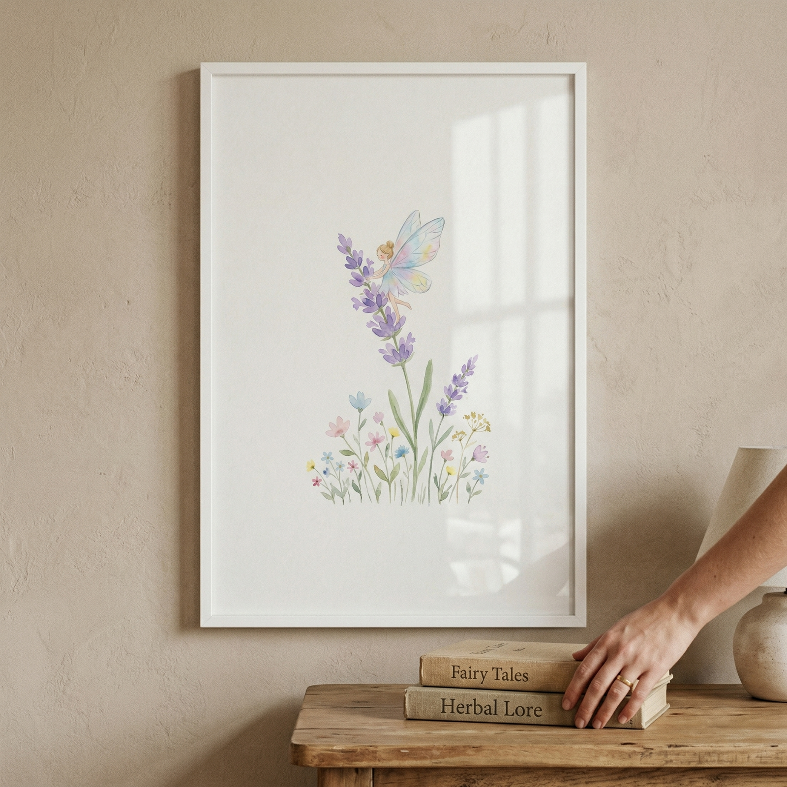 A white-framed art piece hangs on a beige textured wall above a rustic wooden table. The artwork features a delicate watercolor-style illustration. A tiny, whimsical winged figure with translucent wings is shown sitting on a stalk of purple lavender. At the base of the lavender are smaller, colorful wildflowers in light blue, pink, and yellow. On the wooden table below, two vintage-looking books titled "Fairy Tales" and "Herbal Lore" are stacked, with a person's hand resting on them.