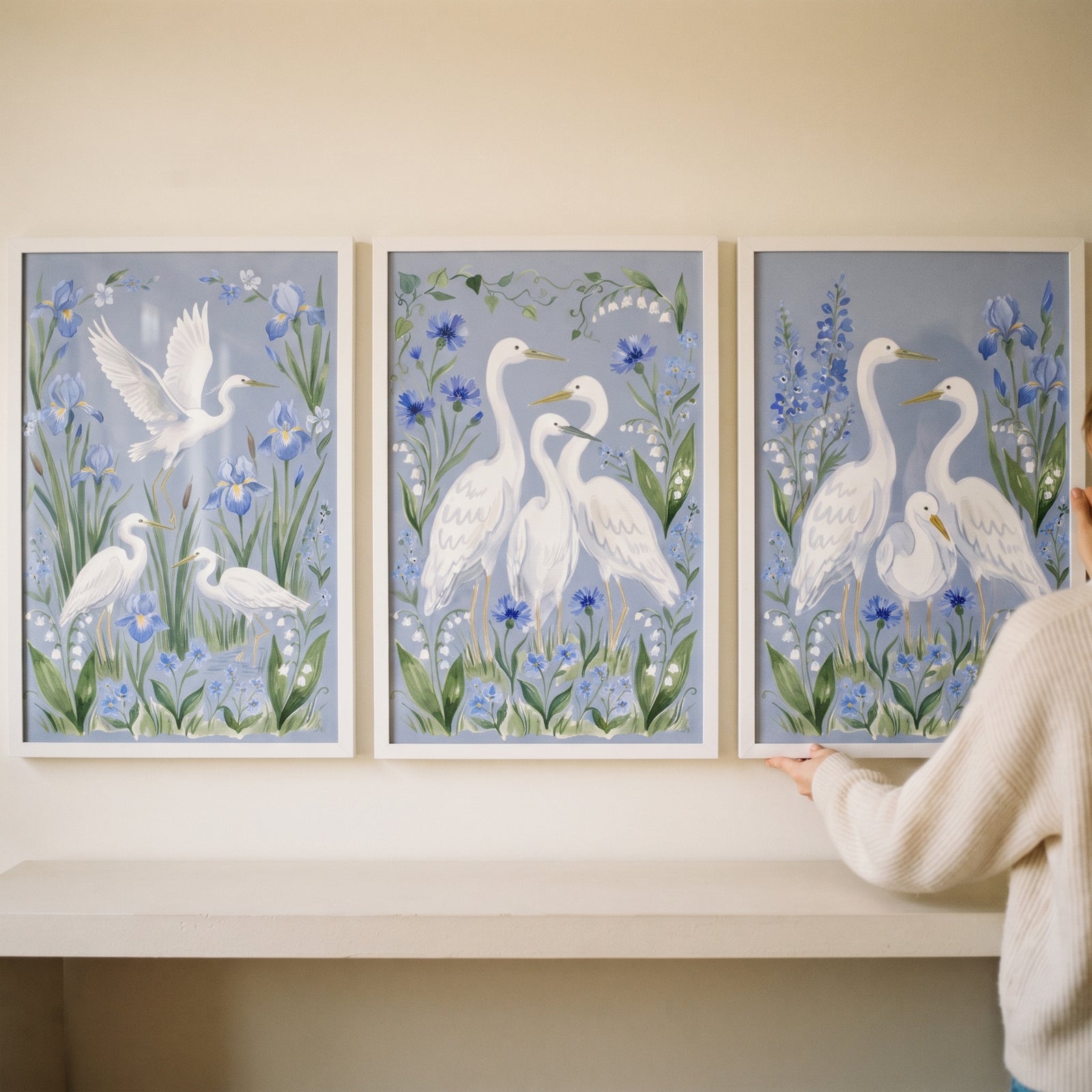 A person hangs a periwinkle floral white egret bird wall art set of 3 featuring white birds and blue botanical flowers.