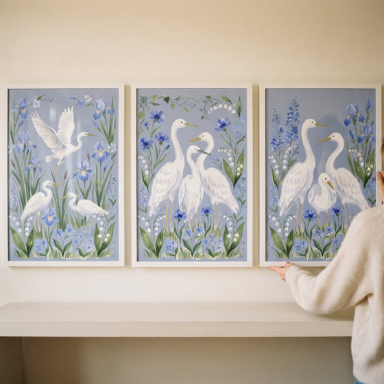 A person hangs a periwinkle floral white egret bird wall art set of 3 featuring white birds and blue botanical flowers.