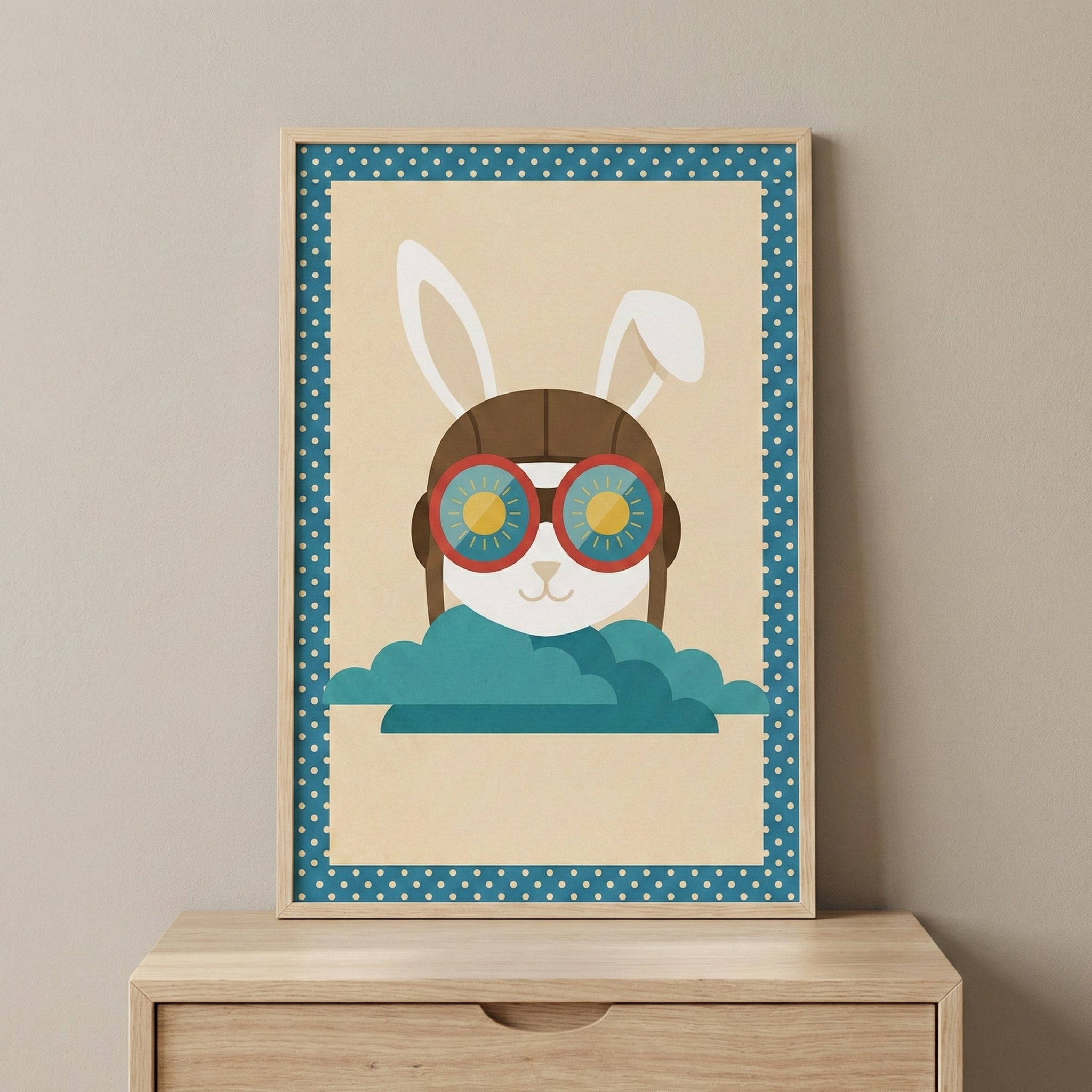 A framed digital illustration of a white rabbit wearing a brown leather aviator cap and oversized red-rimmed goggles, peeking out from behind stylized teal clouds. The rabbit's goggles show a reflection of a yellow sun. The artwork has a light cream background and is framed by a blue border with white polka dots. The piece is housed in a light wood frame and sits on a wooden dresser against a neutral-colored wall.