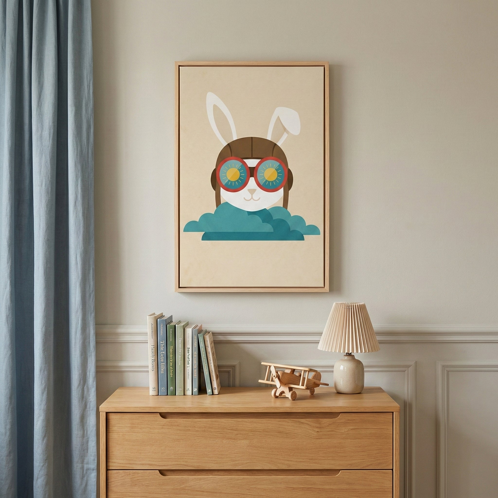 A neutral-toned room featuring a light-colored wooden dresser. On the dresser sits a small row of books, a wooden toy airplane, and a cream-colored lamp with a pleated shade. A blue curtain hangs to the left. Centered on the wall above the dresser is a framed piece of graphic artwork showing a white rabbit wearing a brown aviator cap and large, colorful goggles. The rabbit is depicted peeking out from behind a stylized blue cloud against a light tan background.