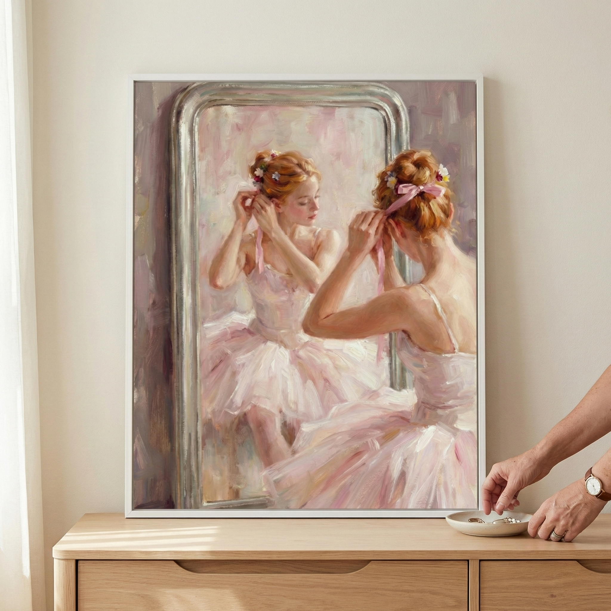 An impressionistic artwork features a ballerina in a soft pink tutu as she prepares in front of a large, silver-framed mirror. Shown from behind, her reflection reveals a gentle expression as she adjusts ribbons and flowers in her hair. The piece is created with light, visible brushstrokes and a delicate palette of pastels. The framed image is displayed on a wooden cabinet, where a person’s hand is visible reaching into a small dish on the surface nearby.