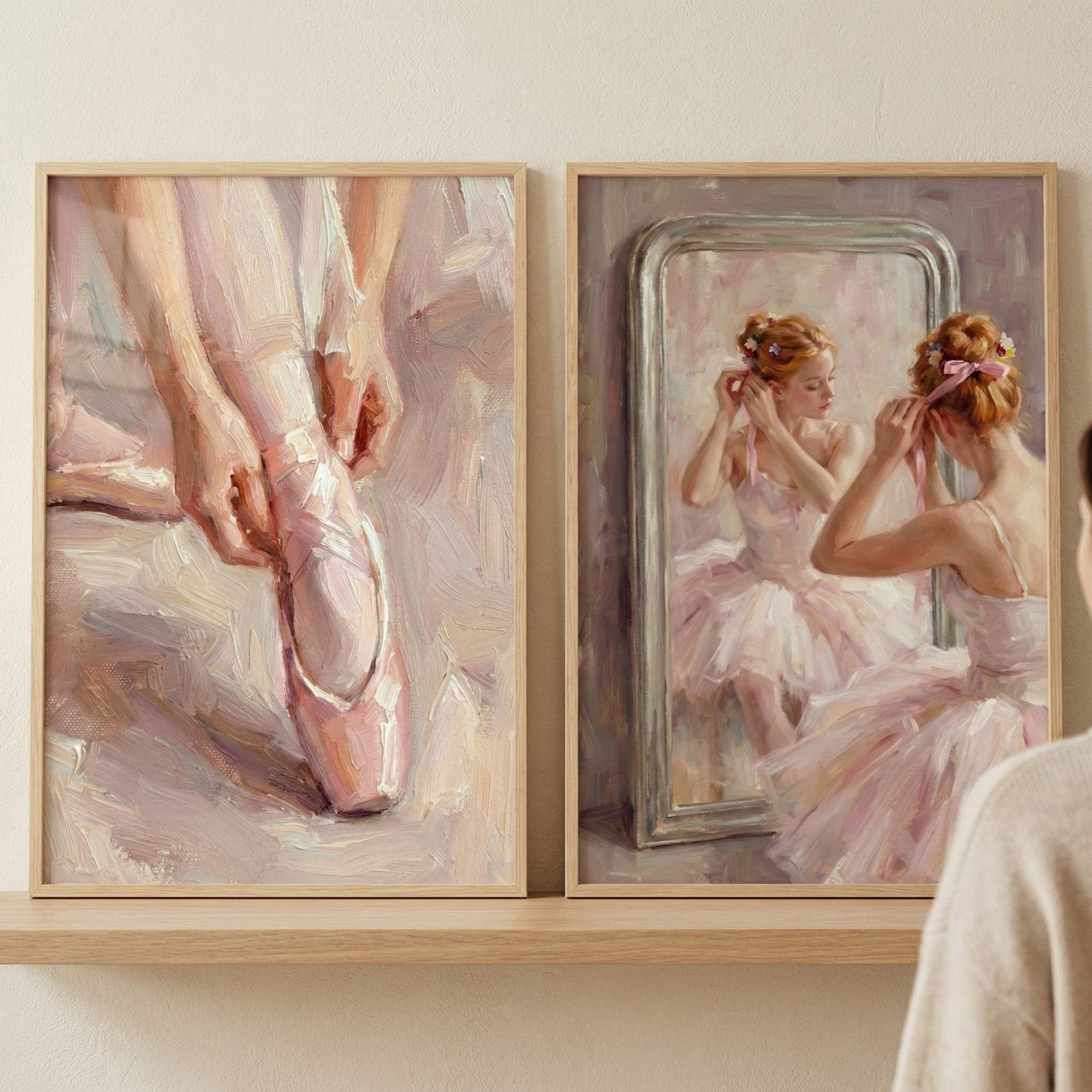 Two light-wood framed artworks sit on a wooden shelf against a neutral wall. Both pieces feature a soft, impressionistic style with visible brushstrokes and pastel colors. The left artwork shows a close-up of a dancer's hands tying the ribbons of a pink pointe shoe. The right one depicts a ballerina in a pink tutu from behind, looking into a silver-framed mirror. Her hair is adorned with flowers and ribbons. A person's shoulder is partially visible in the foreground on the right.