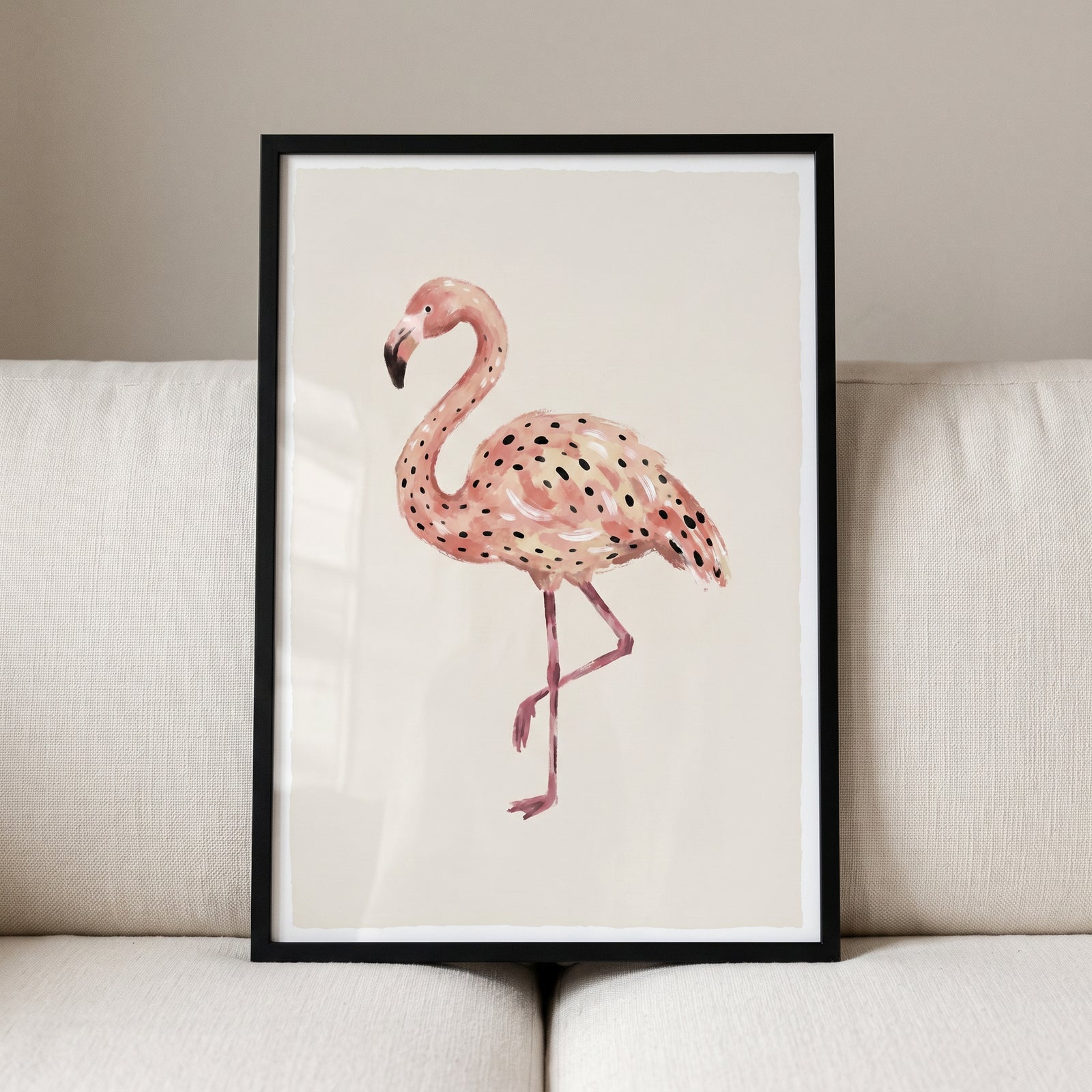 Pink black spotted flamingo wall art in a black frame resting on a cream sofa against a neutral wall.