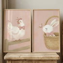 Two white-framed illustrations rest on a minimalist white shelf against a plain, neutral wall. On the left, a charming white chicken stands on a wooden fence, adorned with a pink and white polka-dot bowtie. A butterfly with brown and black patterned wings flutters near its head. On the right, a soft white rabbit sits inside a woven basket, also wearing a matching pink polka-dot bowtie.