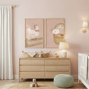 A serene and whimsical nursery room with a soft, pale pink wall. In the center, a light-colored wood dresser with six drawers sits on a light wood floor. Above the dresser hang two light-wood framed illustrations side-by-side. The left frame shows a cute, white cartoon hen wearing a pink bow, perched on a wooden fence. 