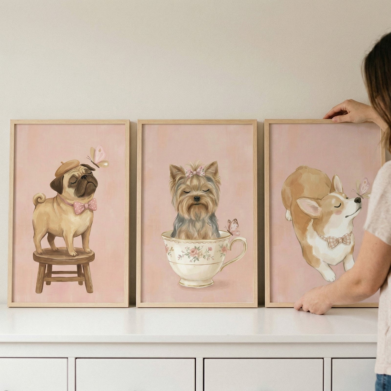 A set of three framed soft-toned illustrations of cute dogs, displayed on a white shelf. Each frame features a different breed against a light pink backdrop. On the left, a small, flat-faced dog with a wrinkled brow wears a tan beret and a pink bow tie while standing on a wooden stool, watching a small pink butterfly. 