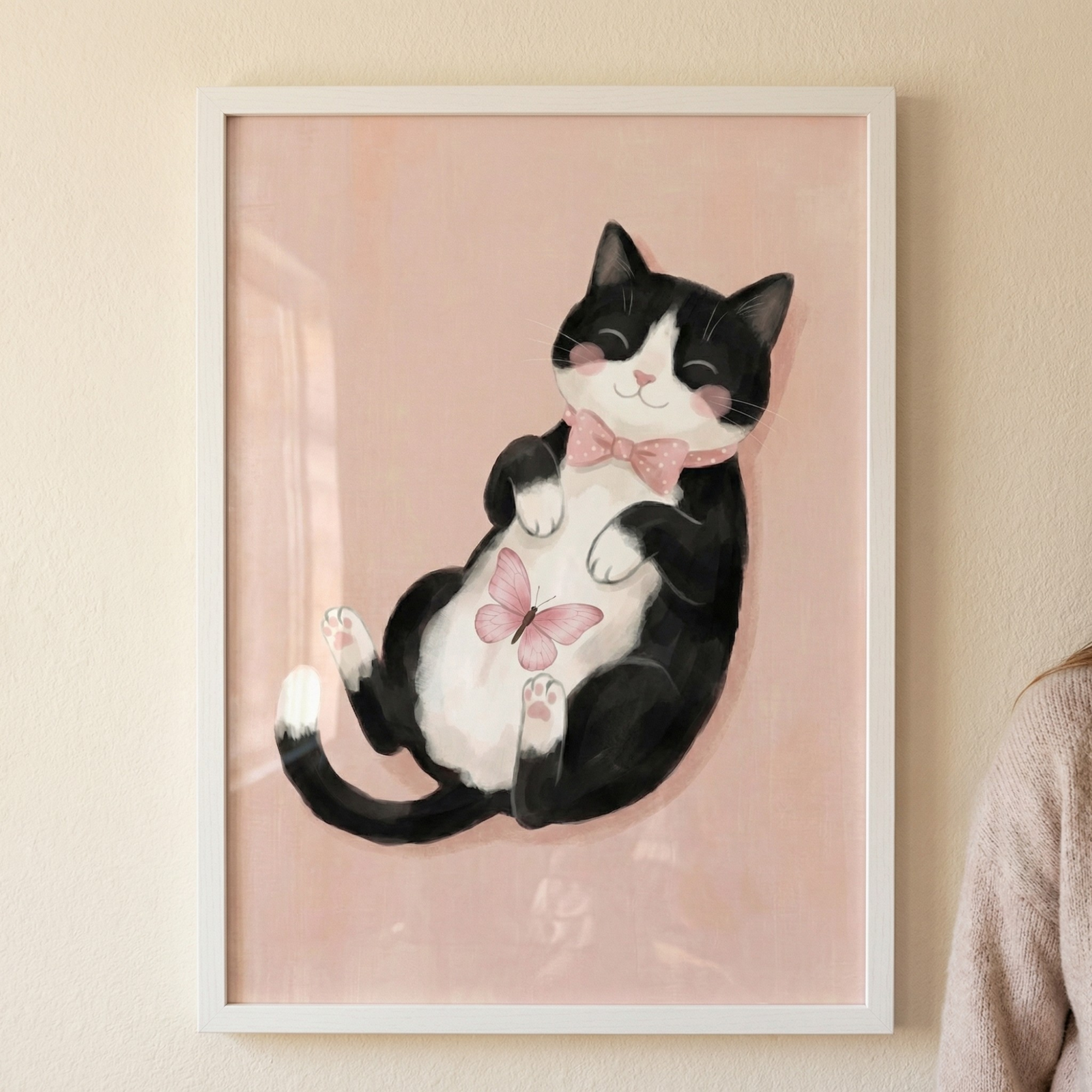 A framed digital illustration hangs on a light-colored wall, featuring a whimsical black and white tuxedo cat lying on its back. The cat has a blissful expression with closed eyes, rosy pink cheeks, and a gentle smile. It wears a soft pink bow tie decorated with white polka dots around its neck. On its white belly, a small pink butterfly is delicately perched. The background of the artwork is a solid, pale pink hue. 