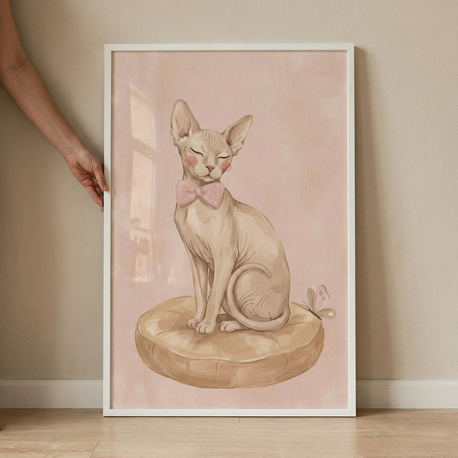 An illustration of a hairless cat sitting elegantly on a tan, tufted cushion. The cat wears a pink bow tie and has soft pink blush on its cheeks, with its eyes peacefully closed. A small butterfly rests on the edge of the cushion. The background is a soft pink wash. The artwork is presented in a slim white frame, which is being held against a beige wall by a person's arm and hand. The bottom of the frame rests on a light wood floor.