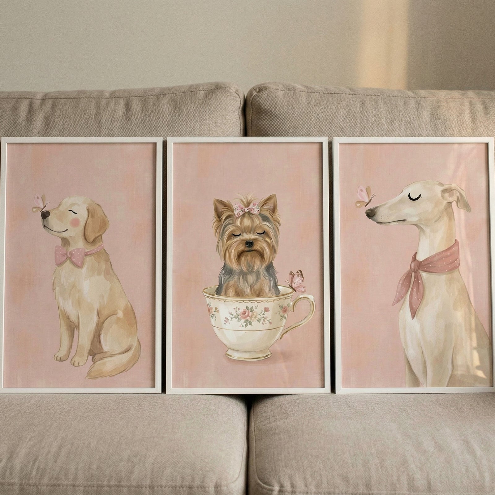 Three framed illustrations are displayed side-by-side against a light-colored fabric sofa. Each piece of art features a different dog against a soft, textured pink background.

The illustration on the left shows a golden-furred dog sitting with its eyes closed in a peaceful expression. It wears a small pink bow around its neck, and a tiny butterfly is perched on its nose.
