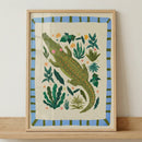 Framed illustration of a green crocodile with pink cheeks surrounded by leafy plants and small colorful flowers in a playful jungle style. The artwork sits on a wooden surface, showing how this whimsical animal print fits into nursery decor.