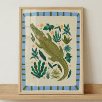Framed illustration of a green crocodile with pink cheeks surrounded by leafy plants and small colorful flowers in a playful jungle style. The artwork sits on a wooden surface, showing how this whimsical animal print fits into nursery decor.