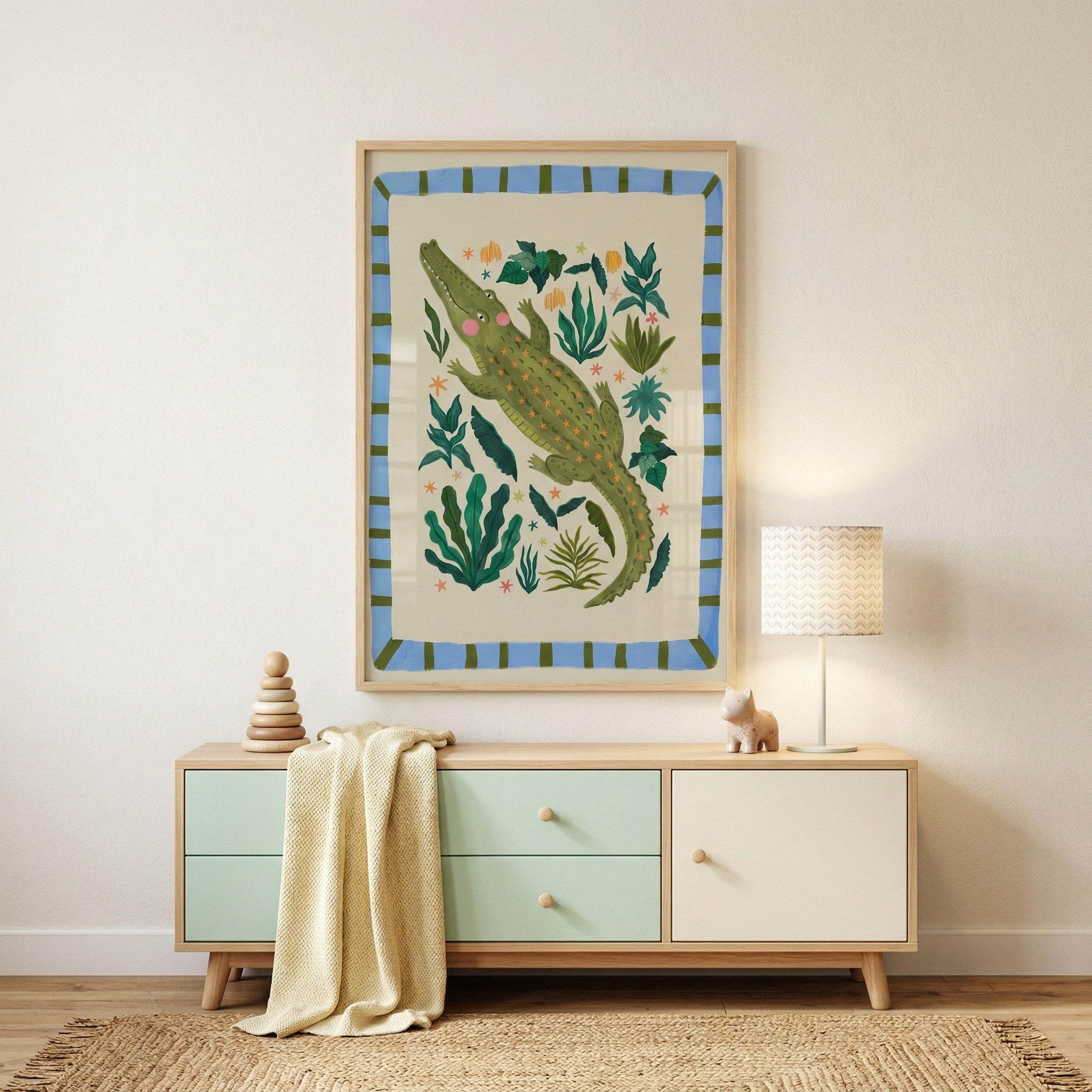 Framed crocodile illustration hung above a light wood dresser with soft green drawers in a calm nursery setting. The playful jungle artwork with leafy details adds color and character alongside a small lamp, wooden toys, and a draped knit blanket.