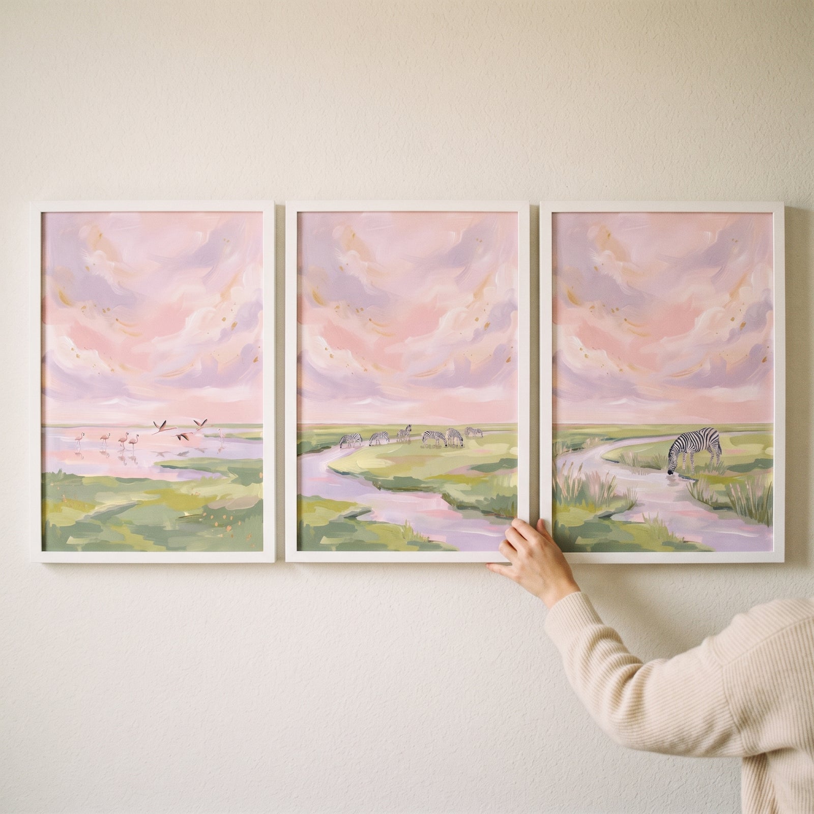 Pink cloudy savanna landscape wall art set of 3 with flamingos and zebras by a river, held against a white wall.