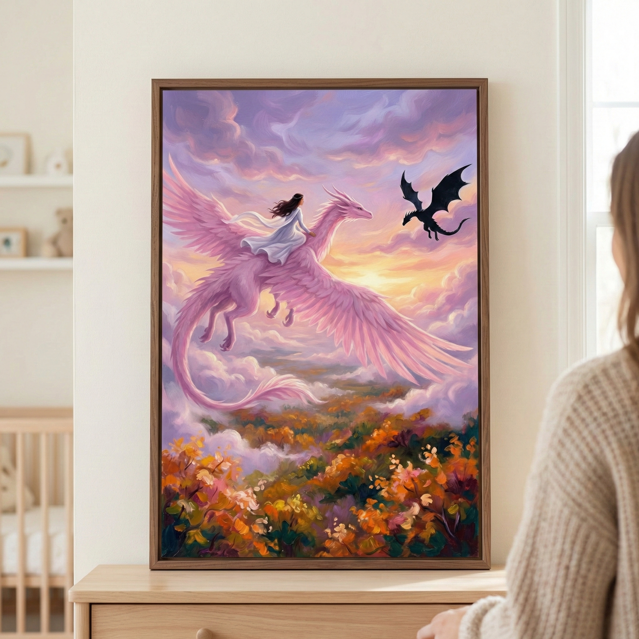 A framed fantasy illustration hangs on a wall, showing a woman in a white dress riding a large, feathered pink dragon through a sunset sky. The sky is filled with soft purple and orange clouds, and a smaller dark dragon silhouette flies in the distance. Below them is a colorful landscape of autumnal trees. A person is partially visible on the right, looking at the artwork, which sits above a wooden dresser. In the background, a child's crib and wall shelves are visible in the soft-lit room.