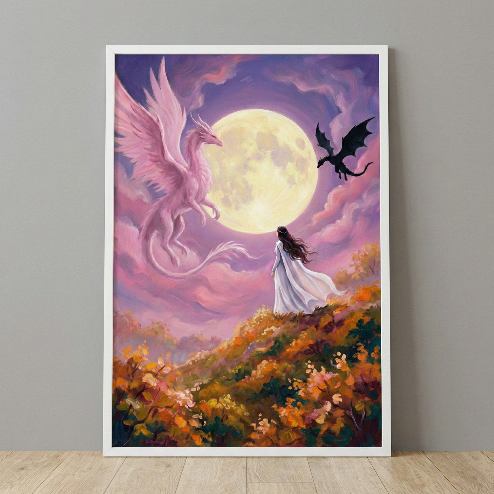 A vertical framed artwork depicts a mystical scene under a large full moon. A woman in a flowing white gown stands on a hillside lush with orange and yellow foliage, gazing upward. To her left, a large, graceful pink dragon with feathered wings glides through purple and pink clouds. On the right, a small black dragon is silhouetted against the bright lunar disc. The illustration is displayed in a simple white frame on a gray wall above a light wood floor.