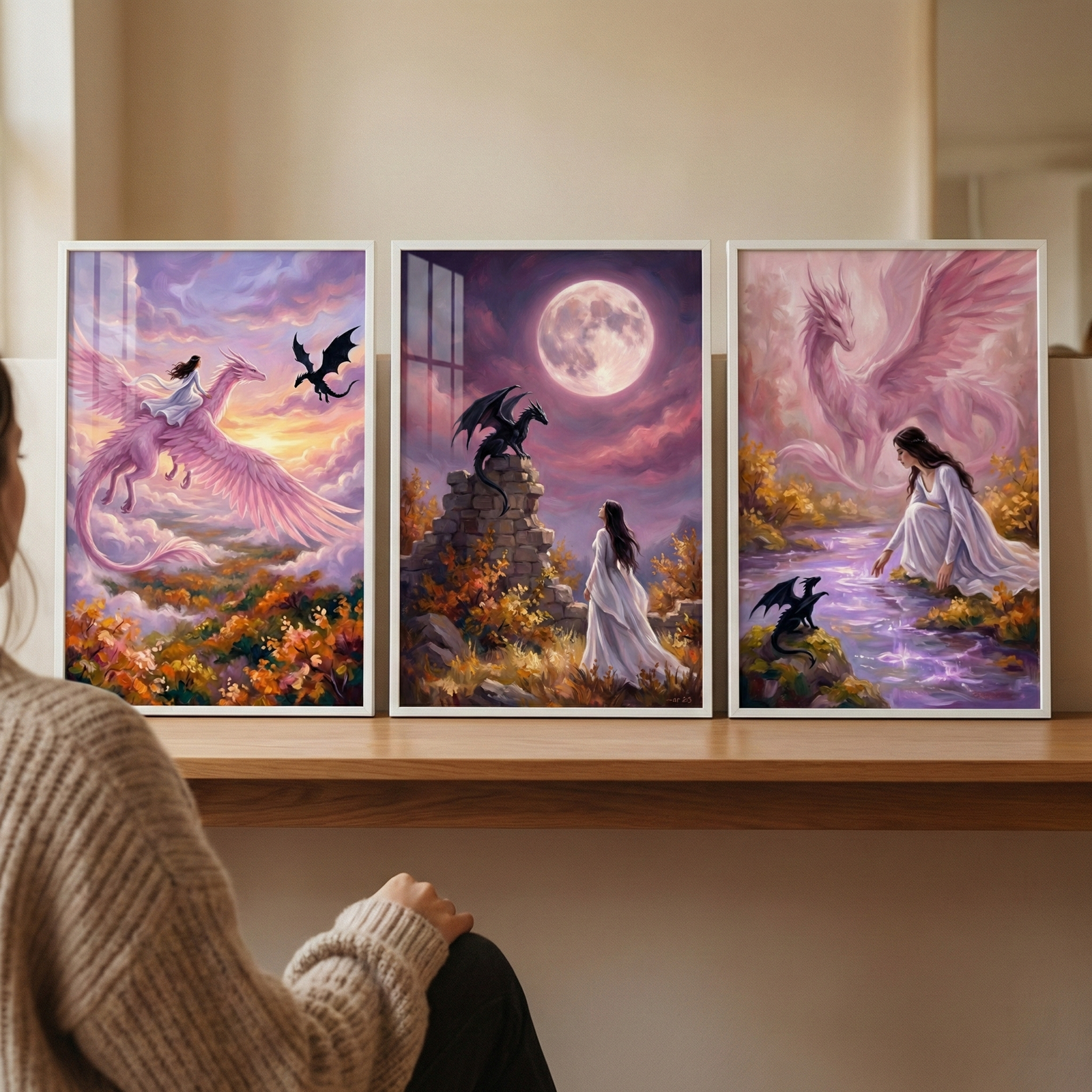 A person in a textured knit sweater sits before three framed artworks displayed on a wooden shelf. The first scene features a woman in a white gown riding a large, pink-feathered dragon across a sunset sky above autumn-colored trees. The middle scene shows the woman in a field, looking up at a large full moon while a black dragon rests on a stone structure. The third scene depicts the woman by a glowing purple river, with an ethereal pink dragon in the clouds and a small black dragon on the bank. 