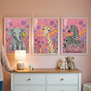 Three framed illustrations featuring cute cartoon animals are displayed on a pink wall in a child's room. From left to right, the frames depict a baby elephant, a giraffe, and two zebras, all set against a pink background filled with colorful wildflowers. Below the frames sits a white dresser with a wooden top. On the dresser, there is a glowing lamp, a small animal figurine, a stuffed bunny, and a teddy bear. A person's arm in a beige sweater is visible on the far left.