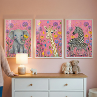 Three framed illustrations featuring cute cartoon animals are displayed on a pink wall in a child's room. From left to right, the frames depict a baby elephant, a giraffe, and two zebras, all set against a pink background filled with colorful wildflowers. Below the frames sits a white dresser with a wooden top. On the dresser, there is a glowing lamp, a small animal figurine, a stuffed bunny, and a teddy bear. A person's arm in a beige sweater is visible on the far left.