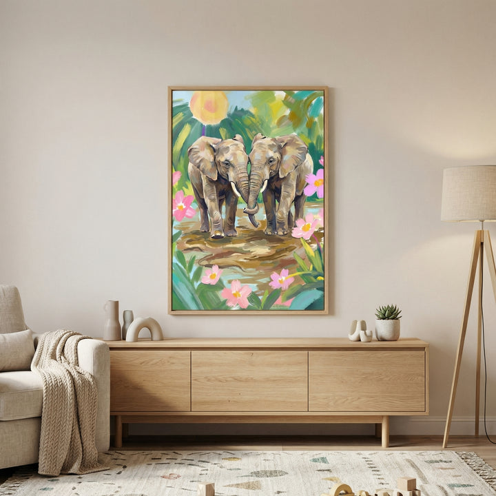 A large, vertically oriented framed illustration hangs on a neutral, light-beige wall in a contemporary living room. The piece depicts two elephants standing close together in a lush, tropical landscape. Their trunks are gently touching, and they stand in a shallow pool of water. 