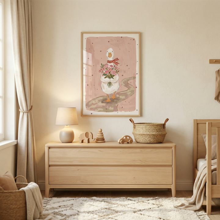 A serene nursery features a light wood dresser centered against a beige wall. Atop the dresser are wooden toys, including an elephant and a stacking ring, a small table lamp, and a woven basket. Above it hangs a framed artwork depicting a white duck with a red scarf carrying a bouquet of pink flowers on a soft pink background. To the right, part of a matching wooden crib is visible. A soft, textured rug with a geometric pattern lies on the floor near a large window draped with neutral curtains.