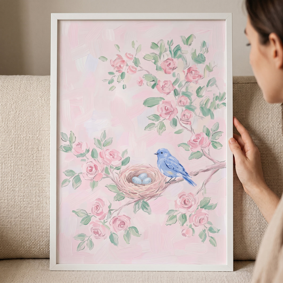 A person holds framed pink floral blue bird wall art featuring a blue bird and nest with eggs among pink roses.