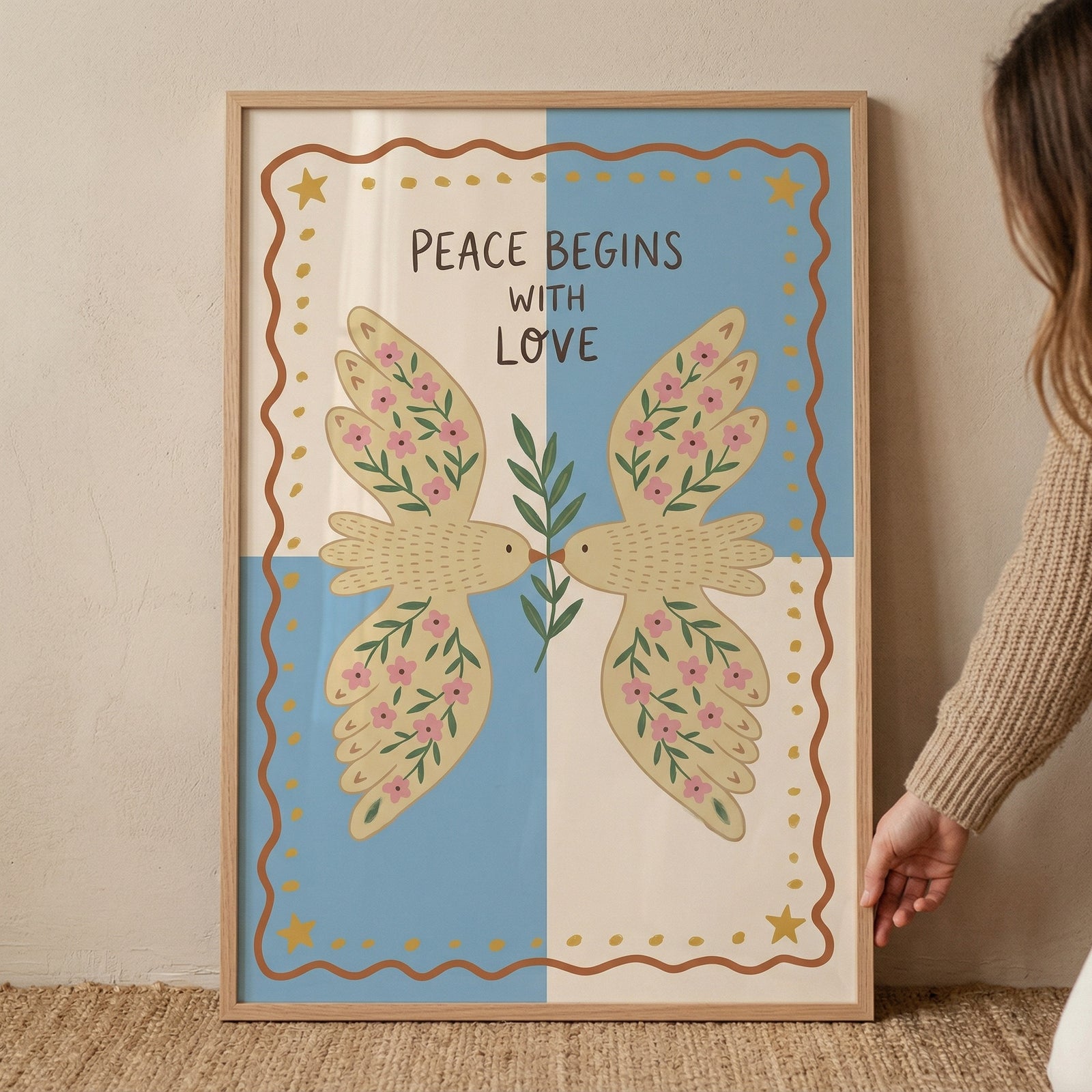 Framed illustration of two birds facing each other holding a green branch, their wings decorated with pink flowers against a soft blue and cream background. The text "PEACE BEGINS WITH LOVE" appears above them, reinforcing a gentle and harmonious theme.