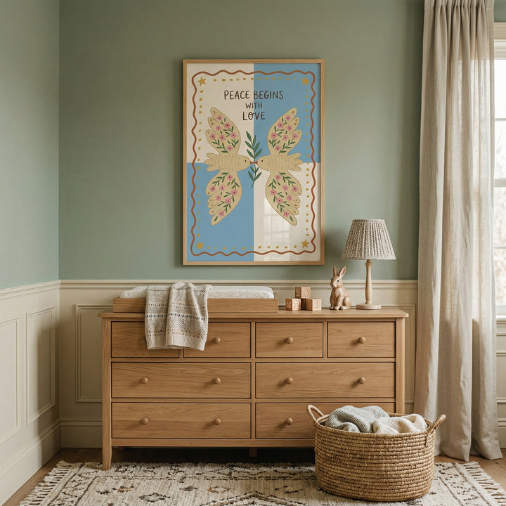 Softly styled nursery with a wooden dresser, woven basket, and neutral decor beneath a framed print of two birds holding a leafy branch. The artwork reads "PEACE BEGINS WITH LOVE" and adds a calm, comforting focal point to the room.