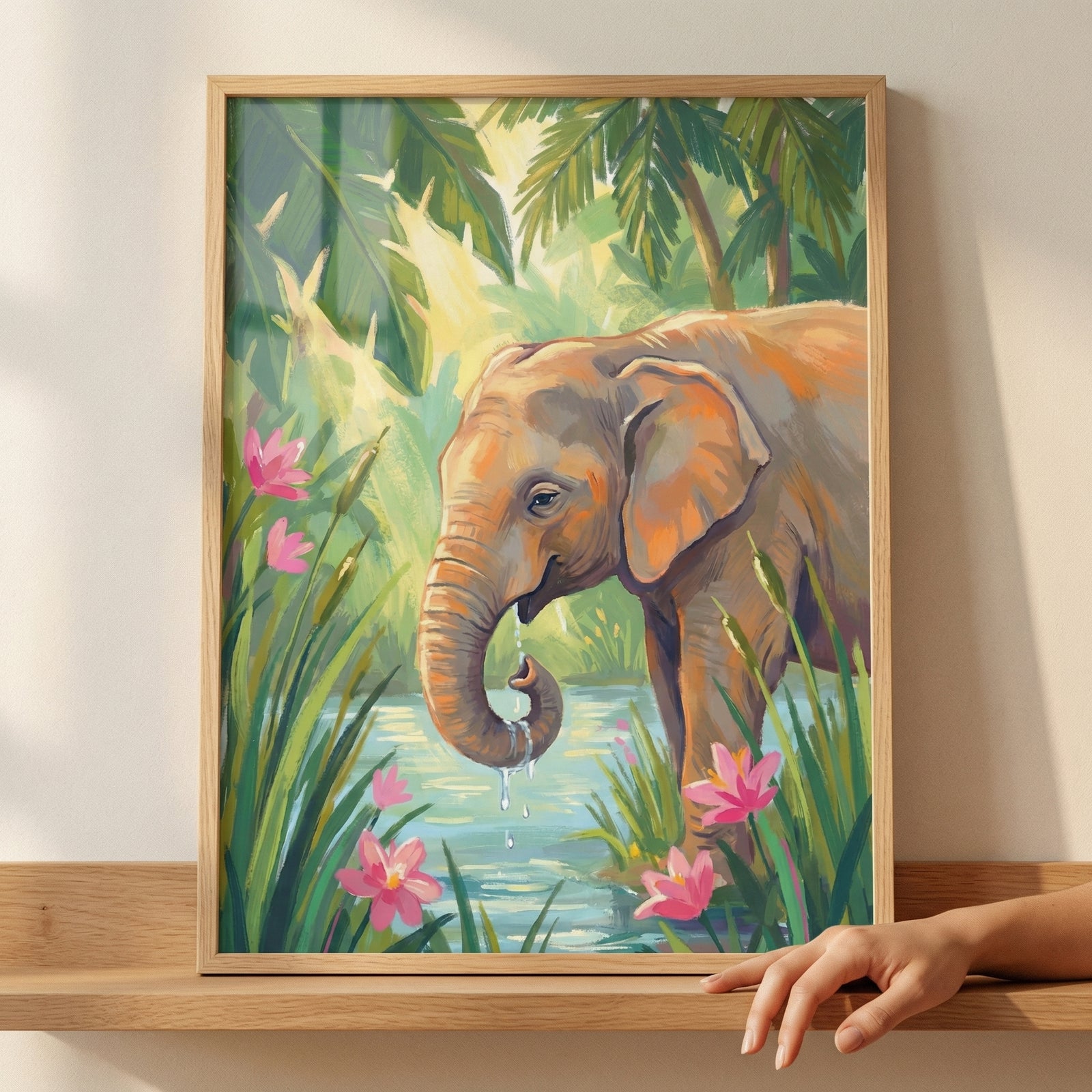 A framed piece of wall art shows a young elephant at a water's edge. The elephant, in warm brown tones, has its trunk curled with water droplets falling from it. The backdrop is a lush tropical jungle with palm fronds. Pink flowers grow in the foreground reeds. The artistic style features expressive, textured brushstrokes and a vibrant palette.
