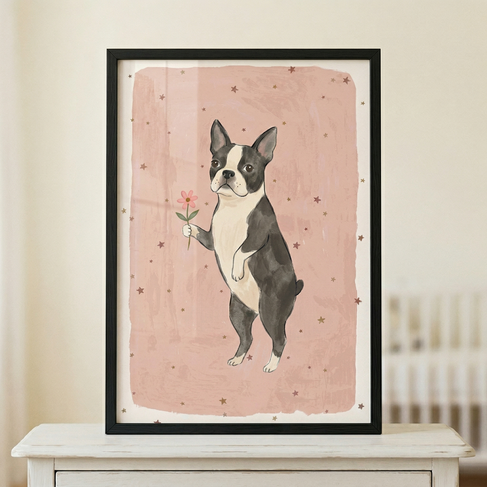 A framed whimsical illustration of a small black and white dog standing on its hind legs. The dog holds a delicate pink flower in its front paw. The background of the artwork is a textured, dusty rose color, peppered with small, simple star shapes. The piece is set in a thin black frame and sits atop a white wooden surface, with a soft-focus background showing part of a room.