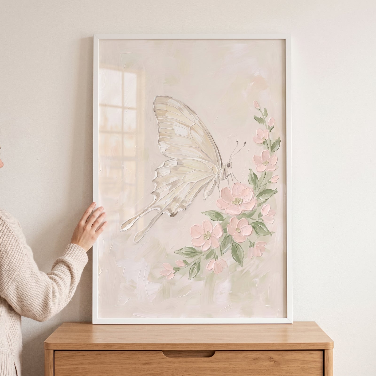 Framed pink flower white butterfly wall art sits on a wooden dresser as a person holds it against a wall.