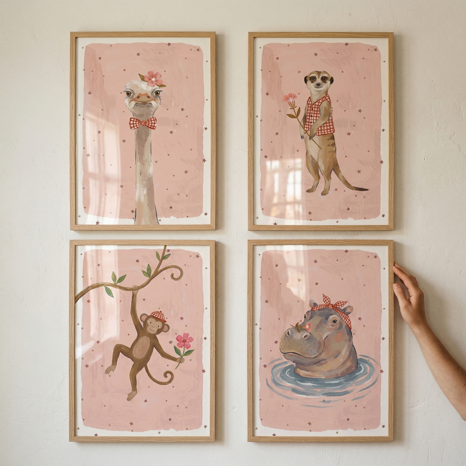 A square grid of four framed animal illustrations is displayed on a light wall. Each artwork features a different animal against a dusty pink background speckled with small stars, with each animal wearing a red-and-white checkered accessory. 