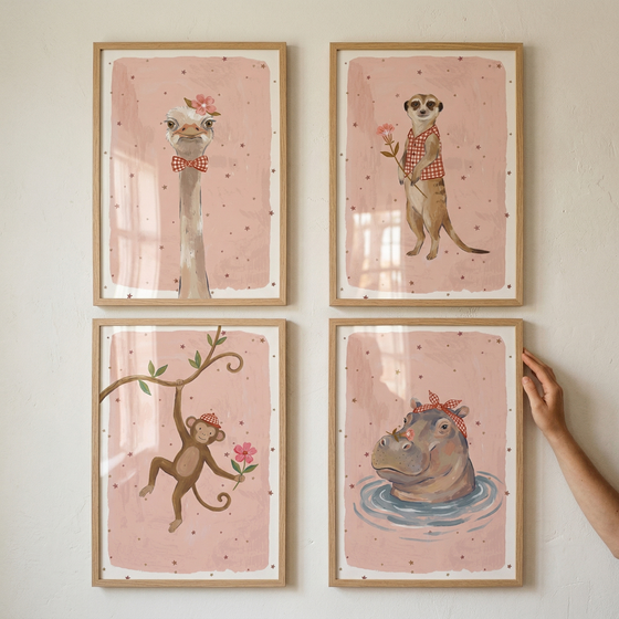 A square grid of four framed animal illustrations is displayed on a light wall. Each artwork features a different animal against a dusty pink background speckled with small stars, with each animal wearing a red-and-white checkered accessory. 