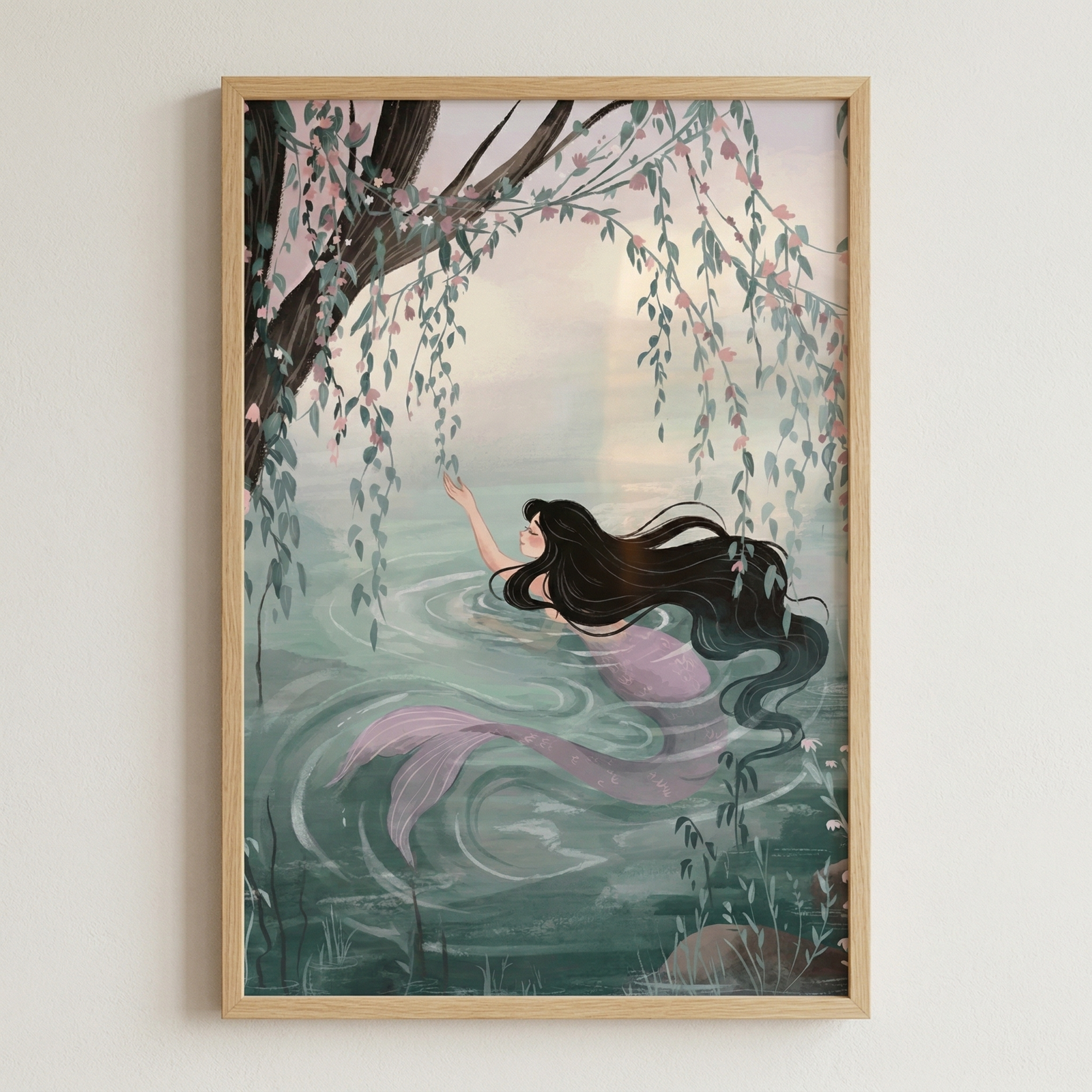 A framed illustration of a mermaid with long, dark hair and a light purple tail swimming in a calm body of water. She reaches up towards the thin, hanging branches of a willow tree decorated with small pink blossoms. The water features soft ripples around her, and the background shows a hazy, pale sky. The entire artwork is presented in a light-colored wooden frame against a neutral white wall.