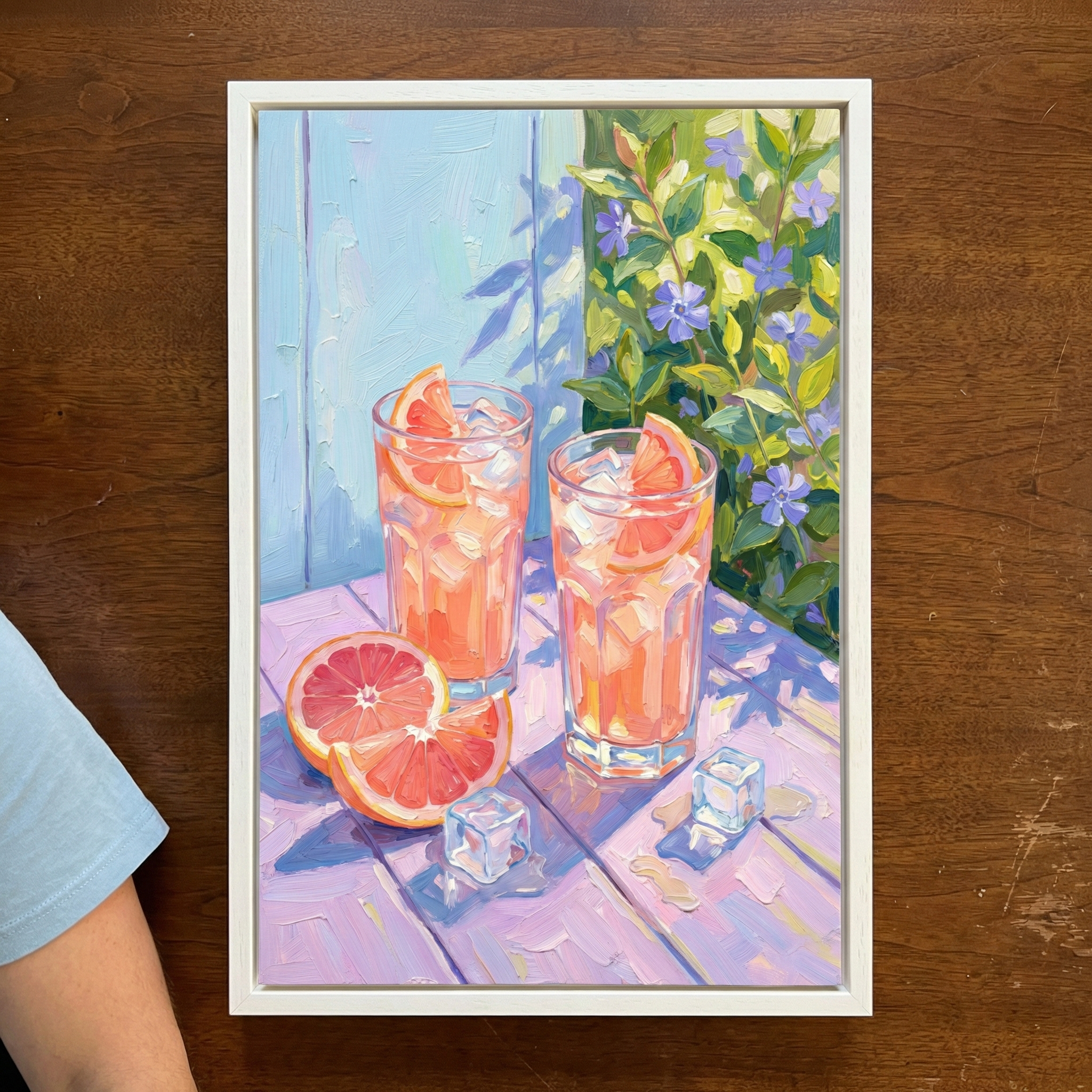 A vibrant still life artwork showcasing two tall glasses of pink citrus drinks with ice. Each is garnished with a grapefruit wedge. On a lavender wooden tabletop, a sliced grapefruit and extra wedges sit near melting ice cubes. The scene is bathed in bright sunlight, casting leaf-shaped shadows onto a light blue wooden backdrop. To the right, green leaves and purple blossoms flourish. The piece features a textured, expressive style with bold brushwork, presented in a white frame.