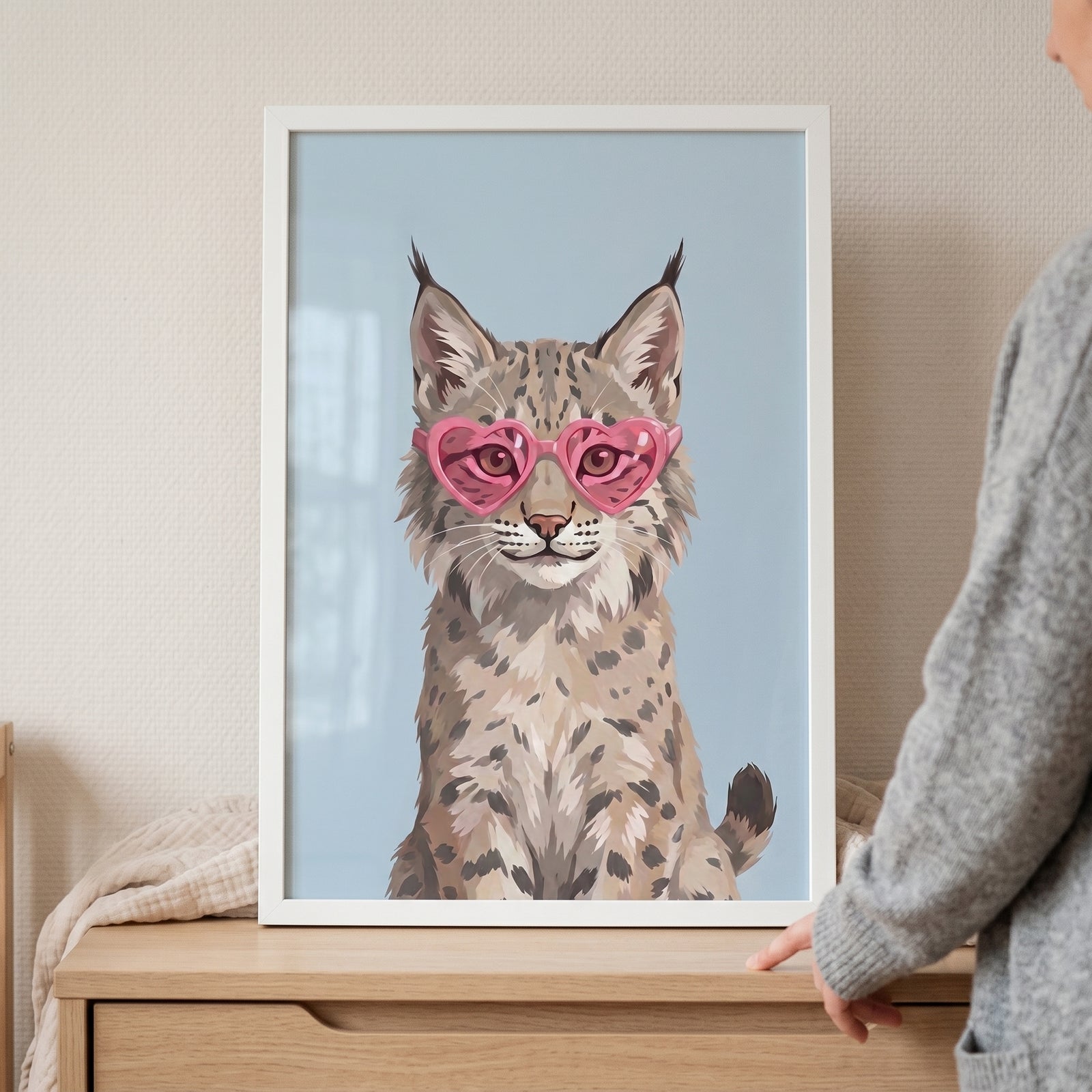 A framed pink heart glasses lynx artwork sits on a wooden dresser next to a person in a grey sweater.
