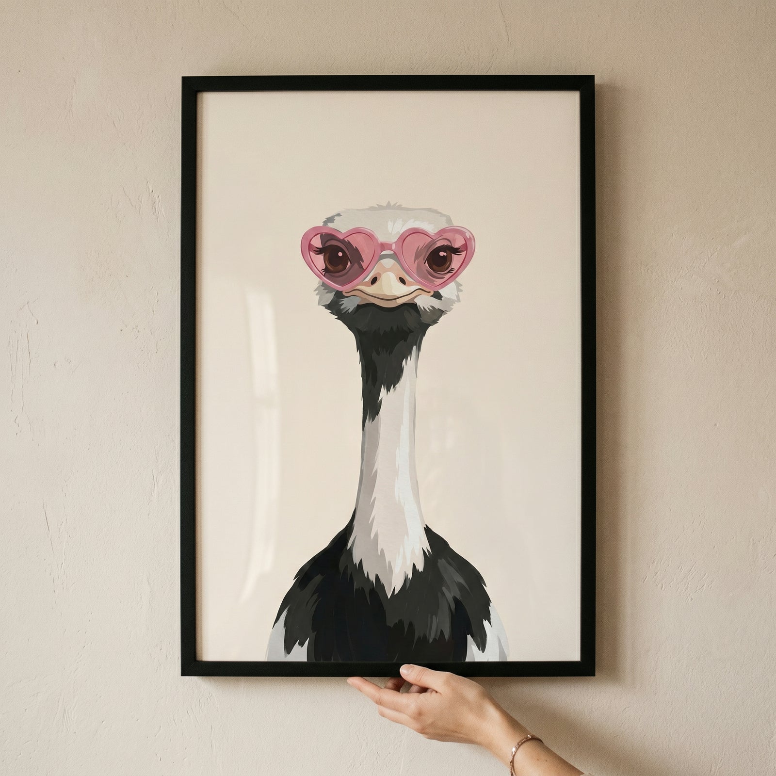 Framed pink heart glasses ostrich wall art held by a hand against a neutral wall.